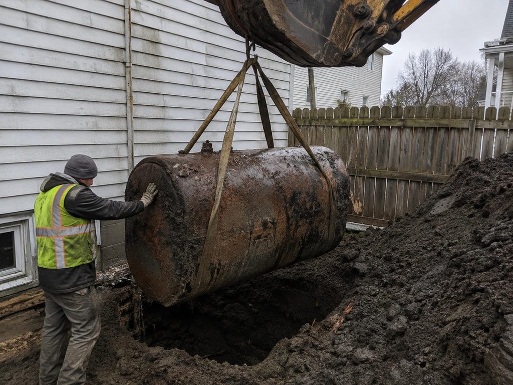 Rusted buried heating oil tank being lifted out of an open excavation in the side yard of an older house