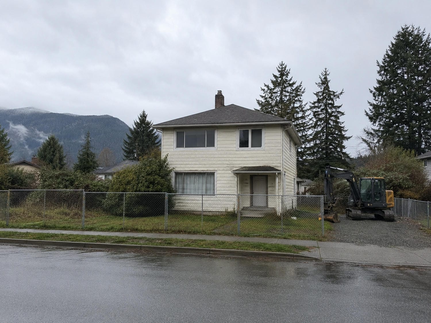 Older wood frame house standing on a fenced Lower Mainland lot with an excavator parked beside it before a custom home teardown