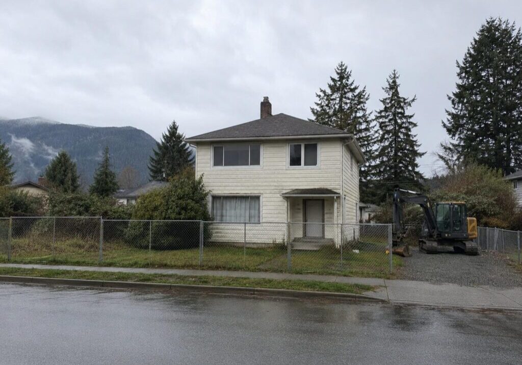 Older wood frame house standing on a fenced Lower Mainland lot with an excavator parked beside it before a custom home teardown